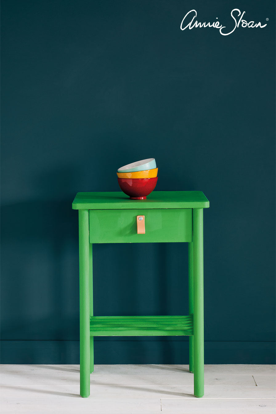 A bright green wooden side table with a single drawer and open lower shelf, styled with a stack of colorful bowls on top, set against a dark teal wall with the Ammie Sloan brand logo in the upper right corner.