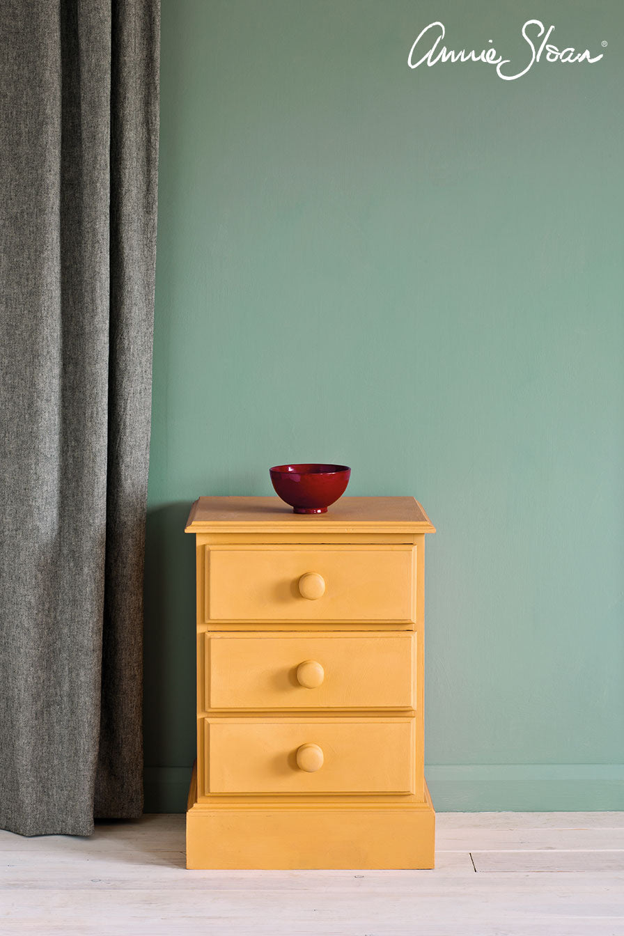 Painted in Annie Sloan Arles Chalk Paint,  mustard yellow three-drawer nightstand with round wooden knobs, set against a sage green wall with a gray curtain on the left. A small red ceramic bowl sits on top.