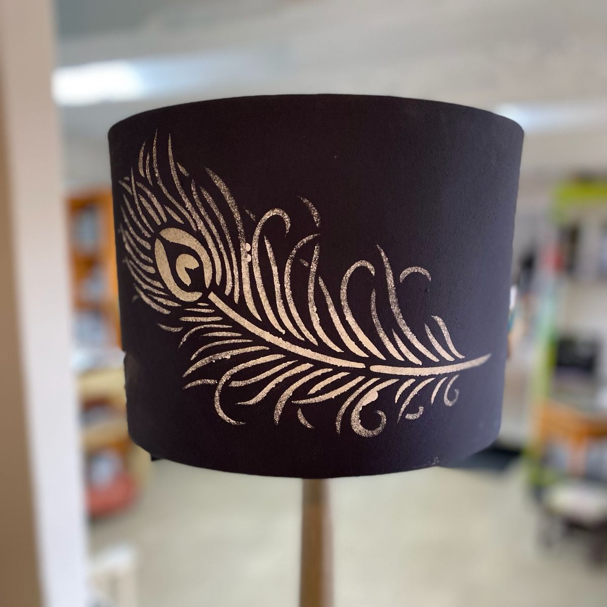 Black lampshade with gold feather design in a blurred indoor setting